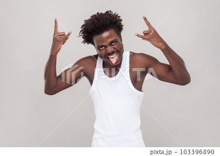 Man with Afro hairstyle shouting from happiness, has widely opened mouth, makes rock n roll gesture, says rock this party. wearing white T-shirt. Indoor studio shot isolated on gray background. 103396890