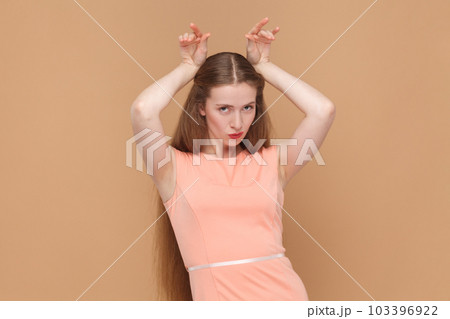 Portrait of funny positive woman with long hair standing with raised arms, showing horns, looking at camera, wearing elegant dress. Indoor studio shot isolated on brown background. Portrait of funny positive woman with long hair standing with raised arms, showing horns, looking at camera, wearing elegant dress. Indoor studio shot isolated on brown background. 103396922