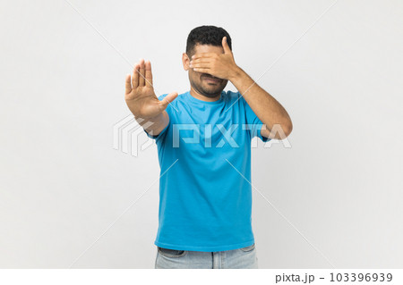 Portrait of young adult unshaven man wearing blue T- shirt standing covering his face with hand, hiding his face, making stop gesture. Indoor studio shot isolated on gray background. 103396939