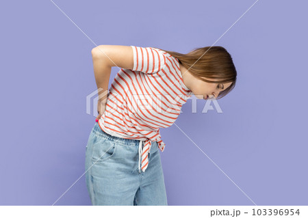 Portrait of unhealthy sick ill blond young adult woman wearing striped T-shirt having discomfort lower lumbar muscular, kidney stones. Indoor studio shot isolated on purple background. 103396954