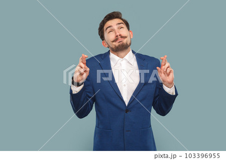 Portrait of hopeful handsome man with mustache standing with crossed fingers, hopes for better and good luck, wearing white shirt and jacket. Indoor studio shot isolated on light blue background. 103396955