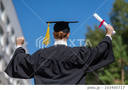 Rear view of a caucasian woman in a graduate gown holding a bundle with a diploma and standing near the university. Rear view of a caucasian woman in a graduate gown holding a bundle with a diploma and standing near the university. 103400717