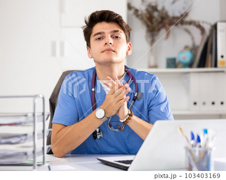 Portrait of man doctor in surgical scrubs sitting at working table in office and using laptop 103403169