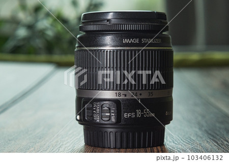 Side view of a DSLR lens. Close-up of the 18-55mm zoom lens standing on a dark wooden background. Green flowers on the back. Side view of a DSLR lens. Close-up of the 18-55mm zoom lens standing on a dark wooden background. Green flowers on the back. 103406132