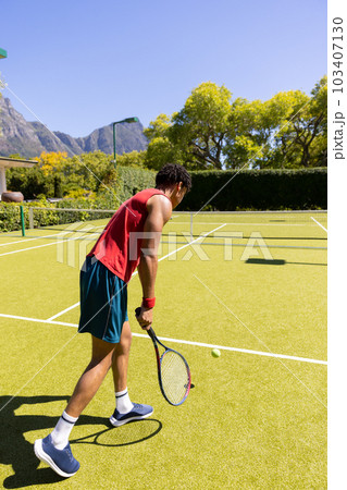Side view of biracial young man serving tennis ball with racket at tennis court against clear sky 103407130