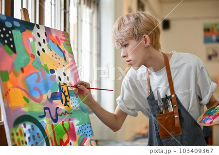 A talented young Asian queer artist focuses on painting his picture on a canvas easel A talented young Asian queer artist focuses on painting his picture on a canvas easel 103409367