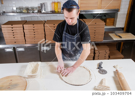 The chef prepares pizza. Raw pizza ready to bake. Cook in a blue apron in the kitchen. with a shovel in his hands. boxes for food delivery on background. The chef prepares pizza. Raw pizza ready to bake. Cook in a blue apron in the kitchen. with a shovel in his hands. boxes for food delivery on background. 103409704