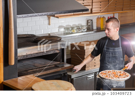 The chef prepares pizza. Raw pizza ready to bake. Cook in a blue apron in the kitchen. with a shovel in his hands. boxes for food delivery on background. The chef prepares pizza. Raw pizza ready to bake. Cook in a blue apron in the kitchen. with a shovel in his hands. boxes for food delivery on background. 103409723