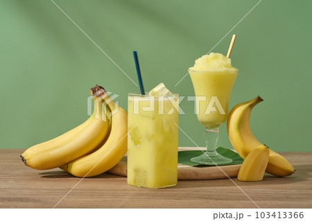 Concept for advertising product with banana ingredient. Ripe bananas and glasses of juice and banana smoothie on a wooden table. Bananas are one of the most consumed fruits in the world Concept for advertising product with banana ingredient. Ripe bananas and glasses of juice and banana smoothie on a wooden table. Bananas are one of the most consumed fruits in the world 103413366