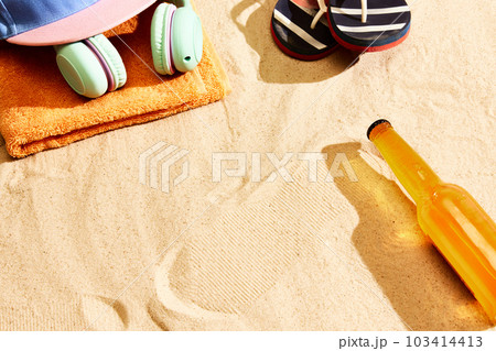 Bottle of lager beer on warm sand with slippers, headphones, towel and cap. Summertime relaxation Bottle of lager beer on warm sand with slippers, headphones, towel and cap. Summertime relaxation 103414413