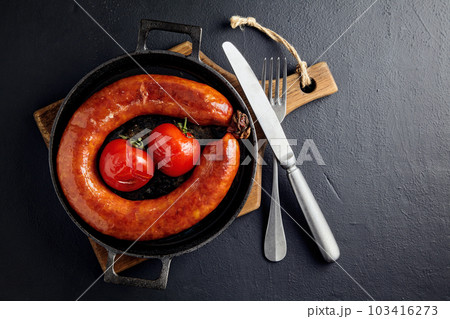 Grilled pork sausage in a cast iron skillet with tomatoes and cutlery on a wooden cutting board on a dark stone table 103416273