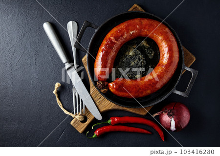 Grilled sausage rolled into a ring on a cast-iron pan with cutlery, a wooden cutting board, hot peppers and red onions on a black stone background Grilled sausage rolled into a ring on a cast-iron pan with cutlery, a wooden cutting board, hot peppers and red onions on a black stone background 103416280