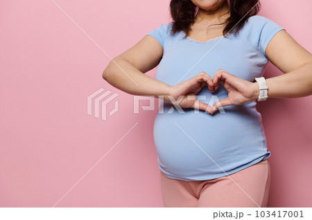 Focus on the hands of a pregnant woman, expectant gravid mother in blue t-shirt, showing heart shape made with her fingers over her big belly, expressing love, tenderness, isolated on pink background Focus on the hands of a pregnant woman, expectant gravid mother in blue t-shirt, showing heart shape made with her fingers over her big belly, expressing love, tenderness, isolated on pink background 103417001