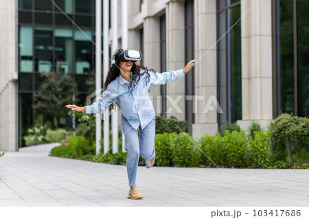 Young beautiful girl in shirt and jeans dancing outside office building wearing virtual reality glasses, hispanic woman happy having fun and relaxing with curly hair. Young beautiful girl in shirt and jeans dancing outside office building wearing virtual reality glasses, hispanic woman happy having fun and relaxing with curly hair. 103417686
