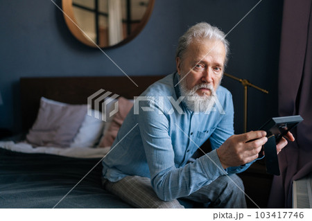 Portrait of unhappy senior adult man stroking framed photo with deep tenderness sitting in dark bedroom, sadness looking at camera. Concept of nostalgia, grief, longing, loneliness in old age. 103417746