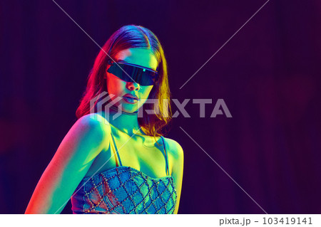 High fashion model wearing futuristic style of clothes with black sunglasses posing over violet studio background in neon light. Close up 103419141
