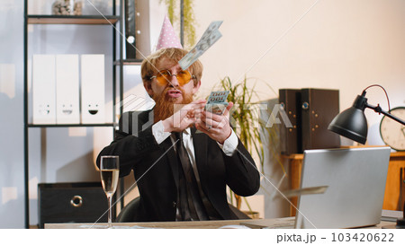 Cheerful rich business man working on office laptop wasting throwing money to camera profit win Cheerful rich business man working on office laptop wasting throwing money to camera profit win 103420622