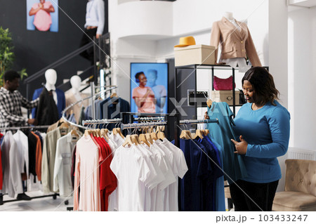 Shopaholic african american woman checking elegant blue shirt, checking formal wear fabric in clothing store. Stylish customer shopping for fashionable merchandise and accessories in modern boutique. 103433247