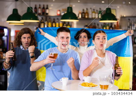 Emotional young adult sports fans waving Ukrainian flag while watching match on TV together in night bar 103434434