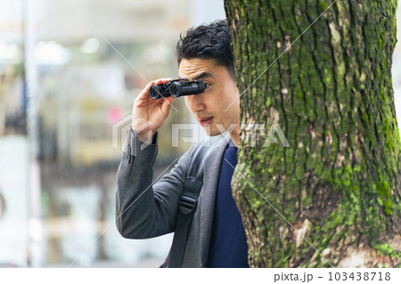 街中で木に隠れて双眼鏡を持って見張りをする男性 103438718
