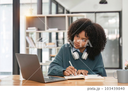 Smiling young african american teen girl wear headphones video calling on laptop. Happy woman student looking at computer screen watching webinar or doing video chat by webcam. 103439840