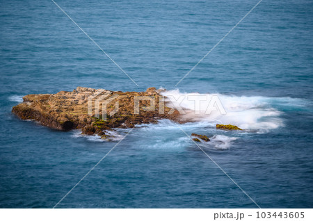 The sea water beats on the brown rocks, producing white spray. 103443605