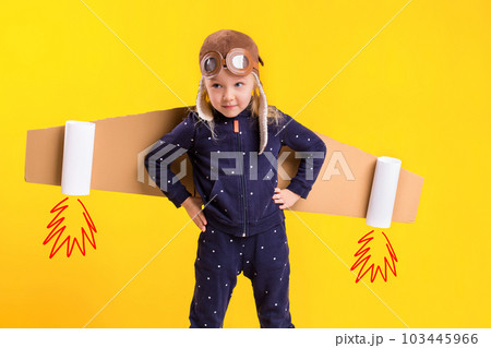 Freedom, girl playing to be airplane pilot, funny little girl with aviator cap and glasses, carries wings made of brown cardboard as an airplane 103445966