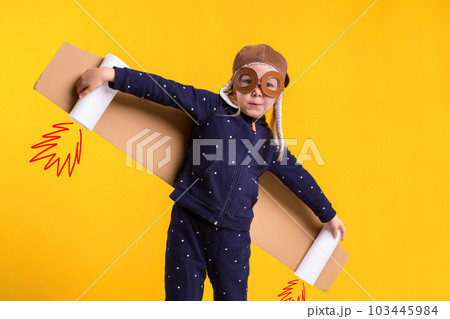 Freedom, girl playing to be airplane pilot, funny little girl with aviator cap and glasses, carries wings made of brown cardboard as an airplane Freedom, girl playing to be airplane pilot, funny little girl with aviator cap and glasses, carries wings made of brown cardboard as an airplane 103445984