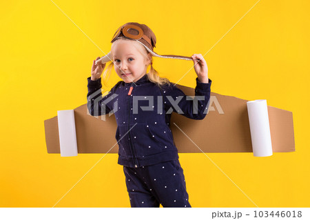 Freedom, girl playing to be airplane pilot, funny little girl with aviator cap and glasses, carries wings made of brown cardboard as an airplane 103446018