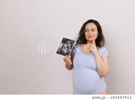 Emotional Latin American pensive pregnant woman, holding ultrasound scan of her baby, dreamily looking aside a copy space on white background. Pregnancy diagnostics concept. Obstetrics and gynecology Emotional Latin American pensive pregnant woman, holding ultrasound scan of her baby, dreamily looking aside a copy space on white background. Pregnancy diagnostics concept. Obstetrics and gynecology 103447413