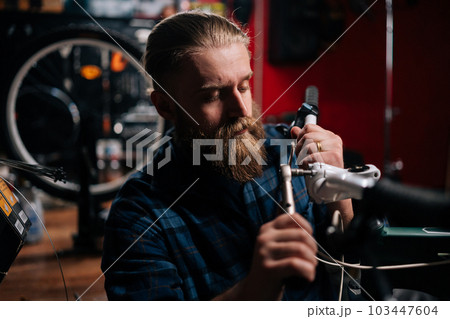 Closeup portrait of concentrated cycling mechanic male checking and repairing bicycle handlebar with special tools working in bike repair shop with dark interior. Concept of maintenance of bike. 103447604