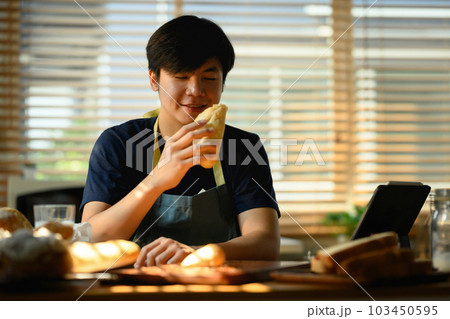 Millennial asian man dressed in casual clothes holding fresh bread and using laptop at dining table in kitchen 103450595