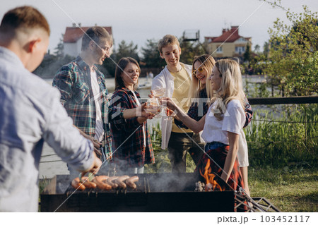 Joyful group of friends is relishing summer vacation with a delightful barbecue. A man grilling sausages over the fire, smoke rises from the meat. Boys and girls are dancing, girl playing the guitar. 103452117