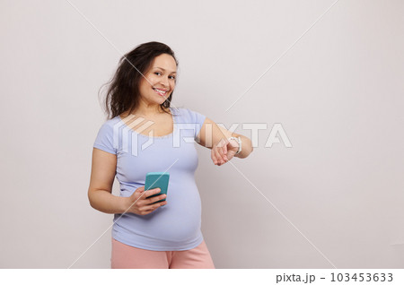 Happy middle aged pregnant woman in 28 week of pregnancy, expecting baby, holding smartphone and checking mobile app on electronic smart wrist watch, smiling looking at camera, over white background 103453633