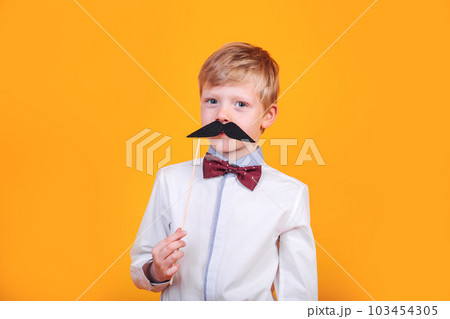 Funny happy hipster kid holding black mustache on yellow background Funny happy hipster kid holding black mustache on yellow background 103454305