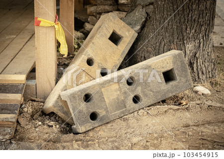 Heavy concrete weight blocks with various holes for temporary construction fence lying near wooden construction and a tree trunk on sand Heavy concrete weight blocks with various holes for temporary construction fence lying near wooden construction and a tree trunk on sand 103454915