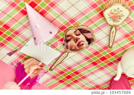 Beautiful young girl with bright makeup looking in mirror lying on checkered tablecloth. Birthday preparation 103457889