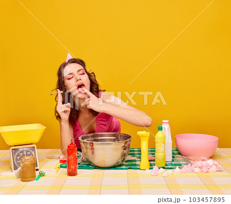 Young beautiful girl with bright makeup eating cake, cooking and smoking against vivid yellow background. Birthday preparation 103457895