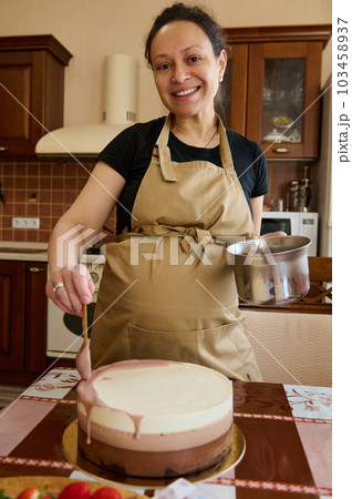 Happy pregnant woman in beige chef's apron, smiling looking at camera, pouring ruby pink chocolate icing over triple chocolate layered mousse dessert with whipped cream and melted Belgian chocolate 103458937
