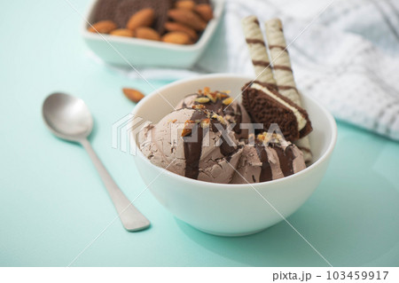 Chocolate ice cream decorated with nuts and chocolate icing, in a white bowl, blue background. Summer homemade cooling desserts Chocolate ice cream decorated with nuts and chocolate icing, in a white bowl, blue background. Summer homemade cooling desserts 103459917