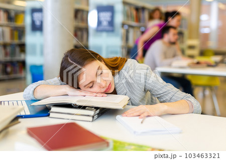 Tired female student sleeping on stack of books in library Tired female student sleeping on stack of books in library 103463321