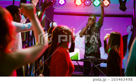 DJ wearing headset on stage at party and mixing music, partying with group of people on dance floor. Young adult feeling happy dancing with friends, using audio equipment. Handheld shot. DJ wearing headset on stage at party and mixing music, partying with group of people on dance floor. Young adult feeling happy dancing with friends, using audio equipment. Handheld shot. 103463498