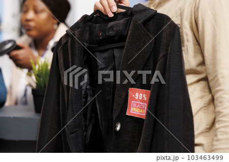 Unrecognizable person customer holding clothes with red Black Friday tag while waiting in line at checkout. People shopping in clothing store during seasonal sales, buying apparel at discounted prices 103463499