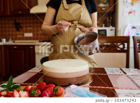 Selective focus on layered delicious cake made from milk, dark and white chocolate - Triple Chocolate mousse dessert on the kitchen table and housewife holding bowl with glaze. Culinary. Confectionery Selective focus on layered delicious cake made from milk, dark and white chocolate - Triple Chocolate mousse dessert on the kitchen table and housewife holding bowl with glaze. Culinary. Confectionery 103473243