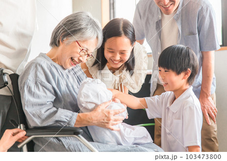 赤ちゃんと対面する車椅子に乗った高齢者女性と子供達・家族 103473800