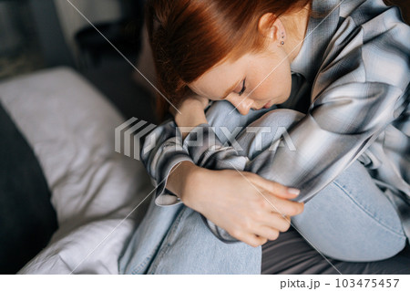 Close-up high-angle view of sad young woman sitting on bed hugging knees lost in bad pessimistic thoughts suffer alone with closed eyes. Stressed female feeling lonely desperate has broken heart. 103475457