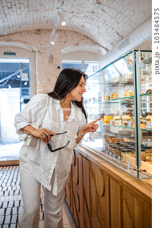 A woman chooses a cake from the window in a coffee shop A woman chooses a cake from the window in a coffee shop 103484575