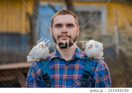A farmer of European Caucasian a man with a beard portrait in a shirt with two dwarf white chickens on his shoulder, close-up against the backdrop of the countryside A farmer of European Caucasian a man with a beard portrait in a shirt with two dwarf white chickens on his shoulder, close-up against the backdrop of the countryside 103489299