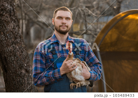 Farmer handsome European appearance male rural portrait on countryside with a beard, shirt and overalls with a white-necked chicken in his arms outdoors Farmer handsome European appearance male rural portrait on countryside with a beard, shirt and overalls with a white-necked chicken in his arms outdoors 103489433