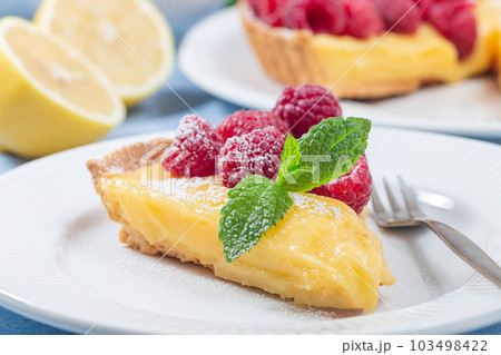 Piece of lemon tart on white plate, garnished with fresh raspberry, icing sugar, mint, horizontal 103498422